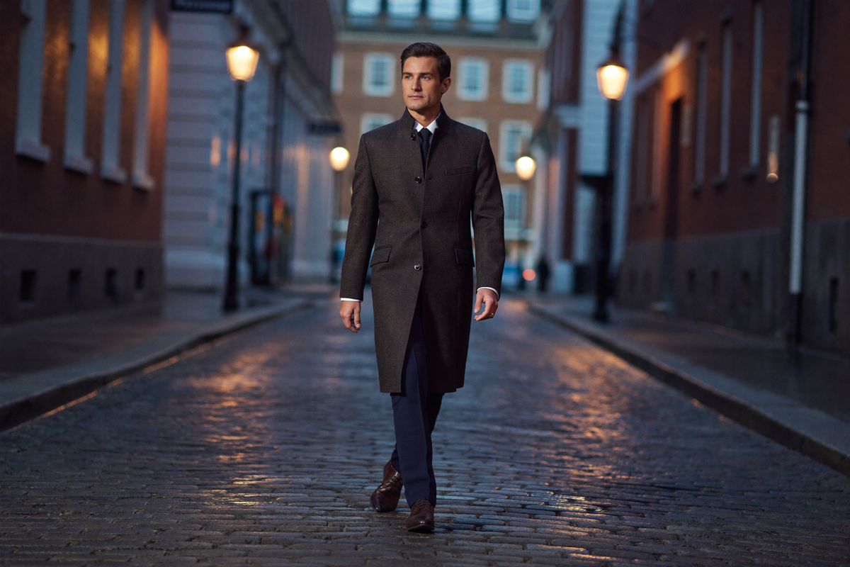 A man in a bespoke charcoal knee-length overcoat walking along a Georgian London street at dusk